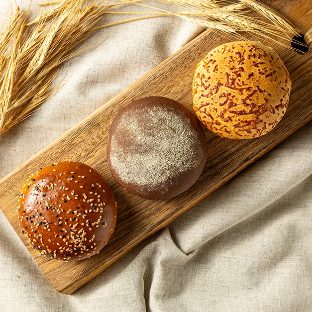 Três pães de hambúrguer artesanais — gergelim, integral e queijo — sobre tábua de madeira